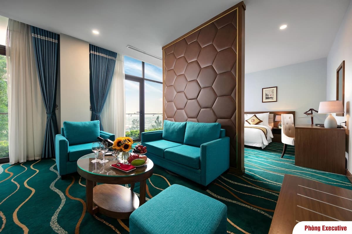 A hotel room with a teal sofa, a round coffee table with a vase of flowers, and a bed with a headboard.