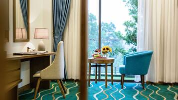A hotel room with a desk, two chairs, a small table with a vase of flowers, and a view of greenery outside.
