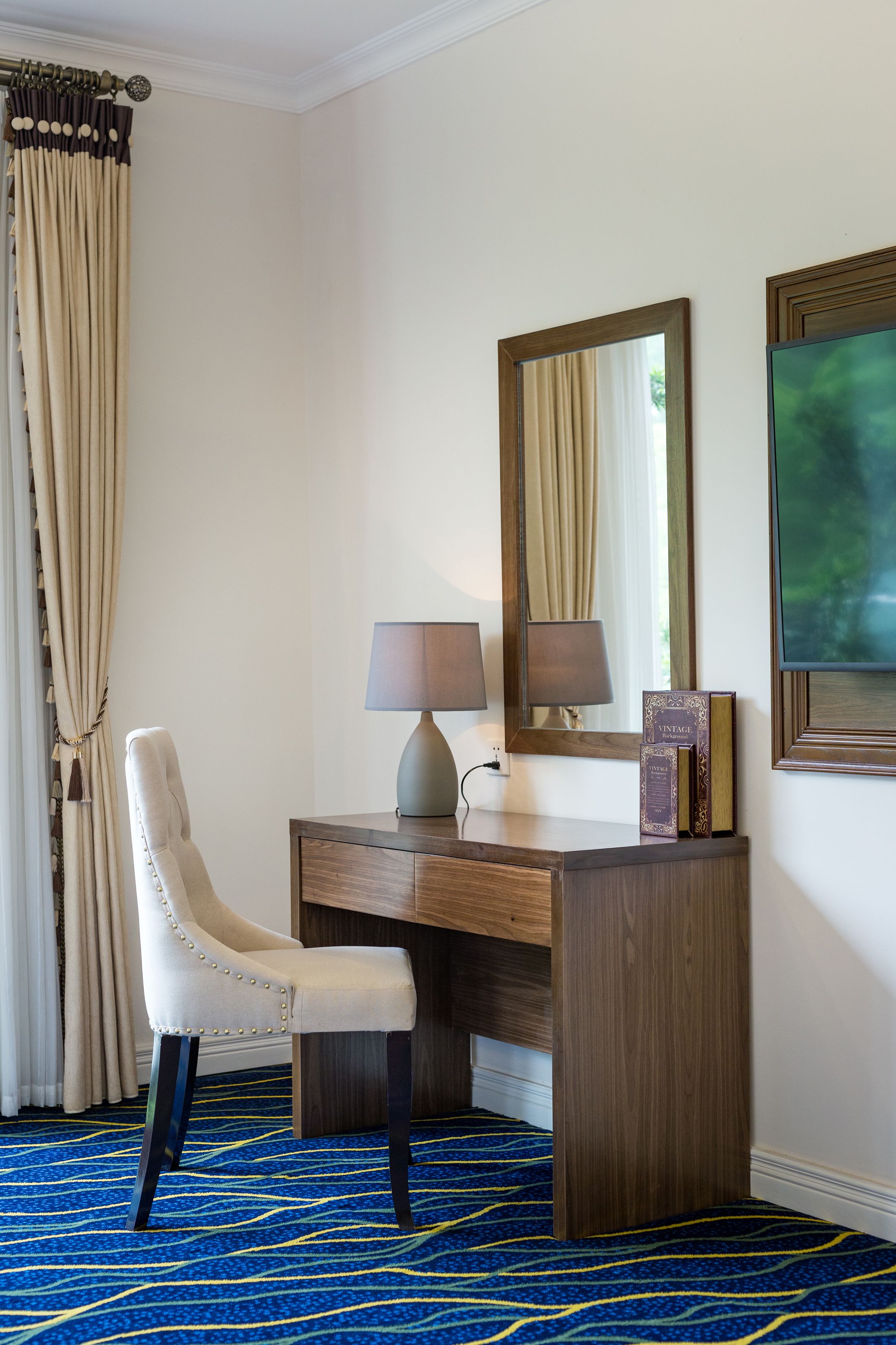 Habitación de hotel con un escritorio de madera, una silla, una lámpara, un espejo y un cuadro enmarcado.