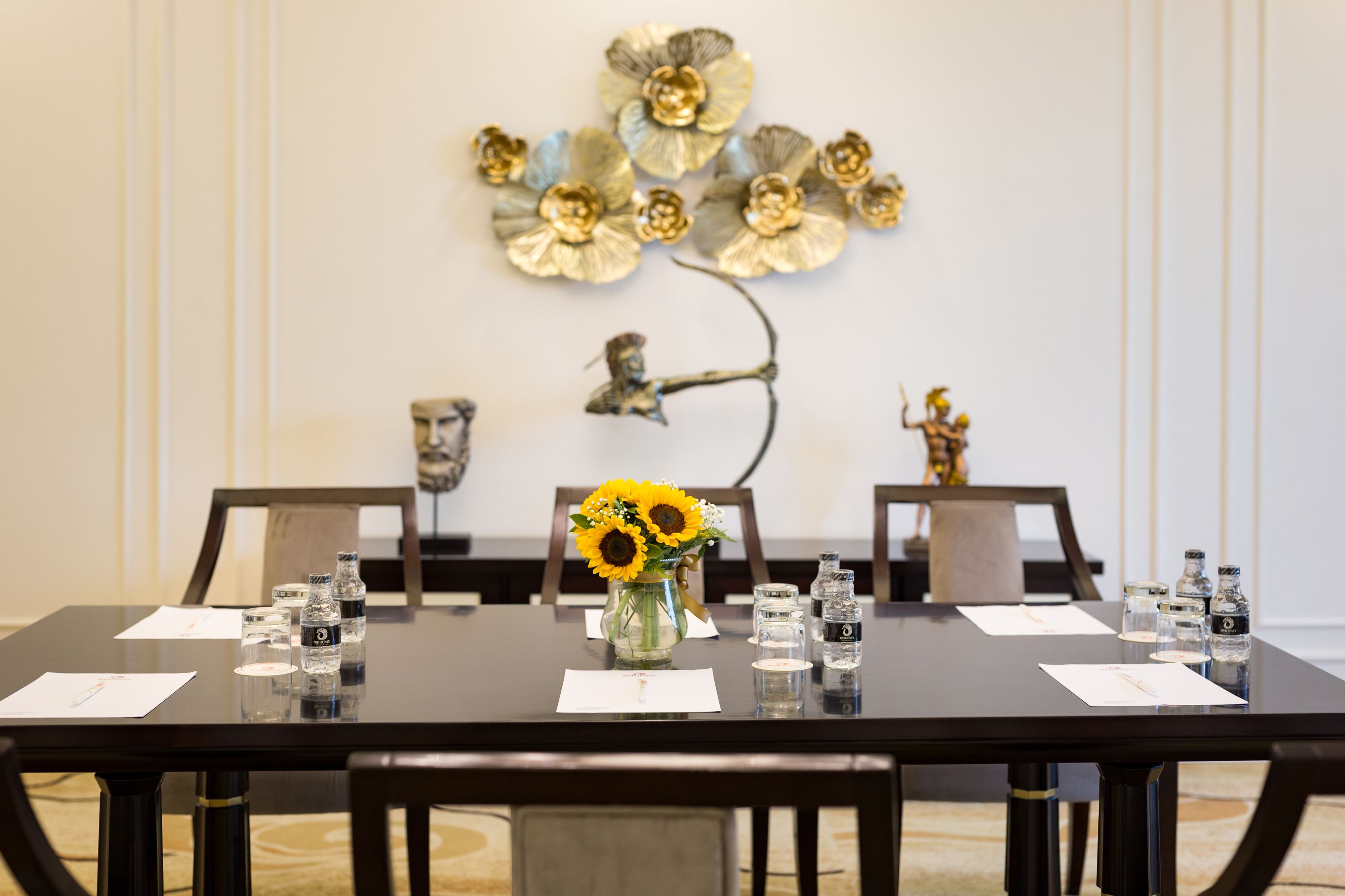 A conference table with water bottles, a vase of sunflowers, and a decorative metal wall piece.