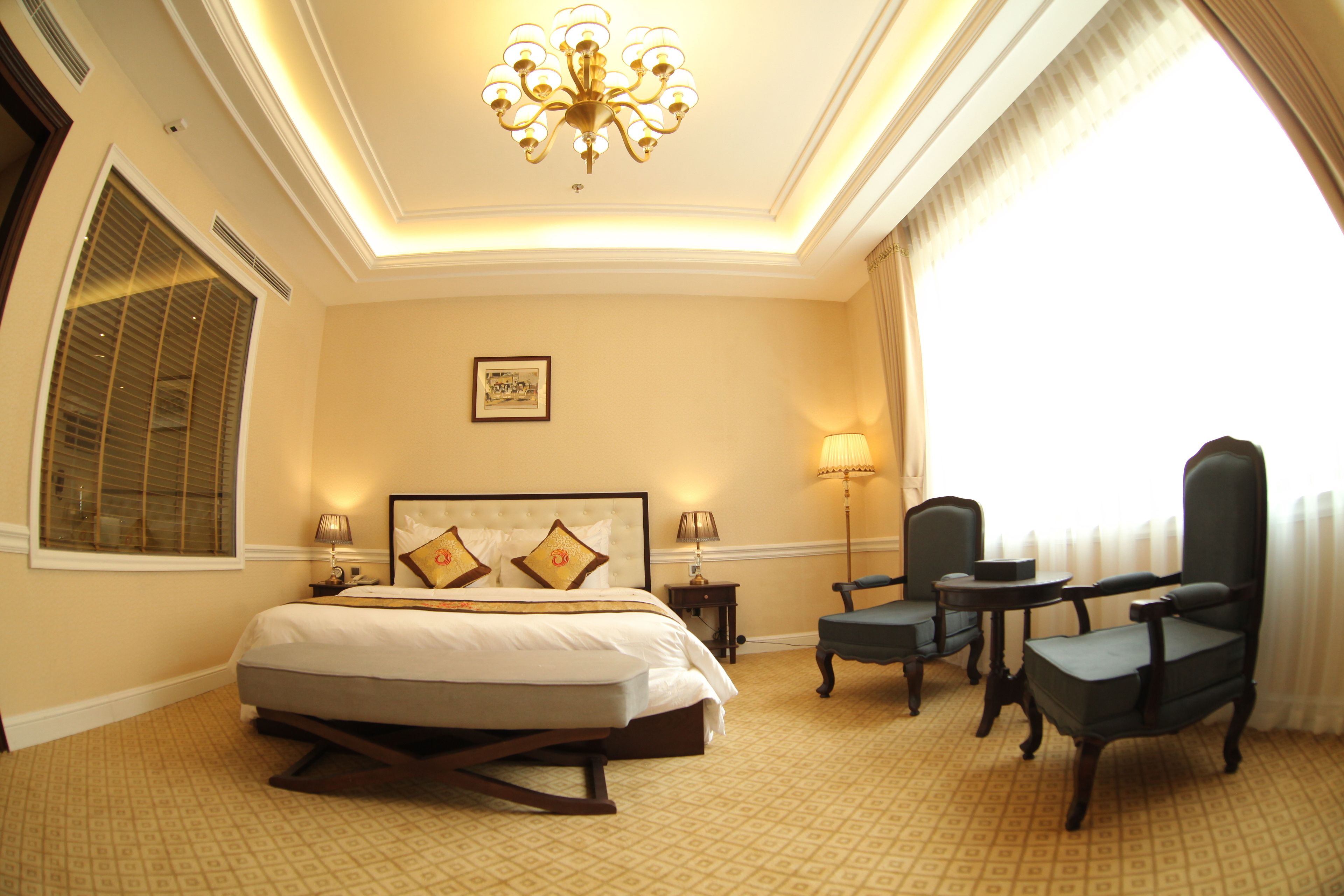 A hotel room with a large bed, two chairs, a small table, a chandelier, and a window with curtains.