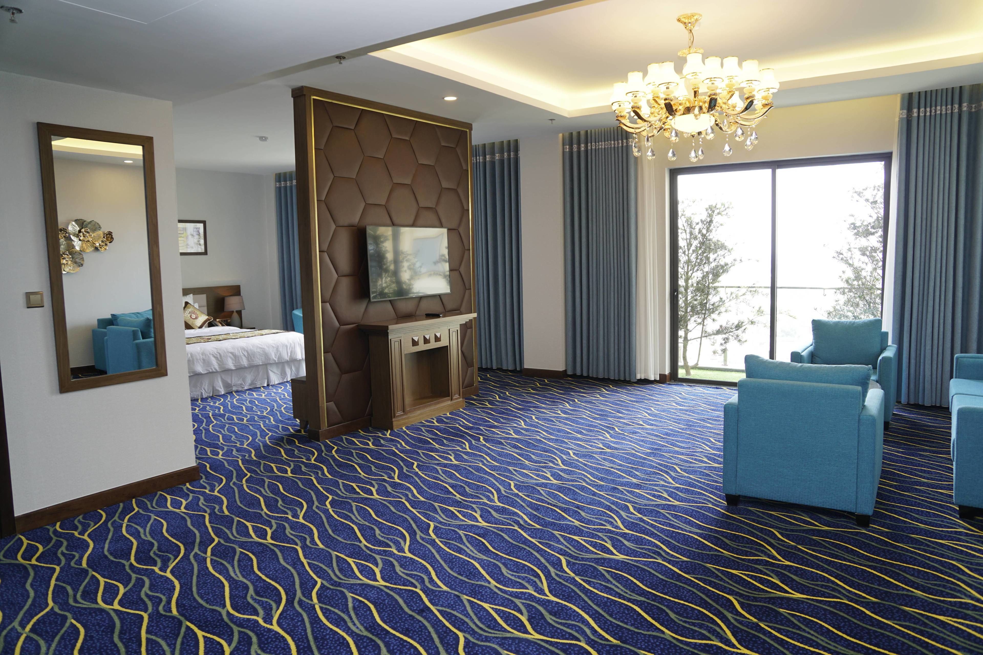 Habitación de hotel con una cama grande, un televisor instalado en un panel de madera, un candelabro y un ventanal con cortinas.