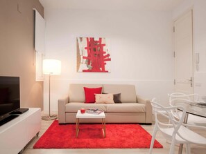 A modern living room with a beige sofa, red and grey cushions, a small white table with a red rug, a TV on a white cabinet, and a glass table with white chairs.