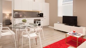 A modern kitchen with white cabinets, a microwave, and a coffee maker. There is a dining area with a glass table and white chairs. A TV is on a white stand, and a red rug is on the floor. - Stylish apartment in city center B1 (Barcelona)