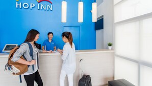Un'area di accoglienza con una parete blu, una scrivania della reception e un cartello con la scritta “HOP INN”. Tre persone stanno interagendo alla scrivania, e si vede una valigia nera a destra.