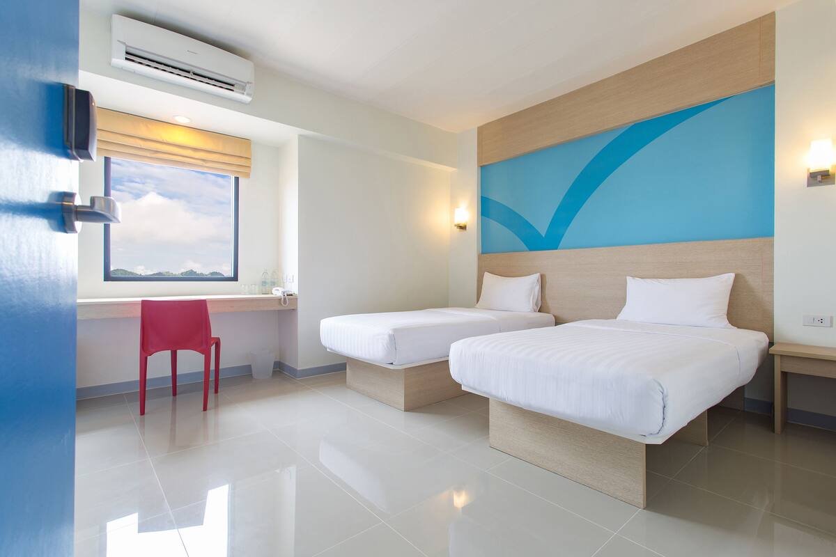 A hotel room with two beds, a red chair, a desk, and a large window with a view.