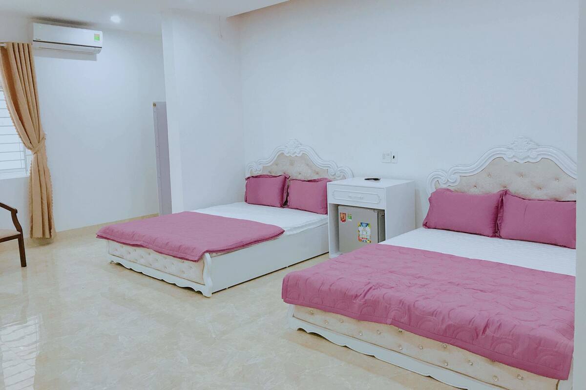 Habitación de hotel con dos camas, cada una con cabecero blanco y ropa de cama rosa. Se observa una pequeña mesita de noche blanca con refrigerador, y una ventana con cortinas color beige.