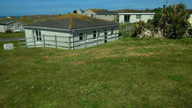A single-story building with a grey exterior and a sloped roof, surrounded by a grassy area and a wooden fence. - F51 The Gables, Riviere Towans (Hayle)