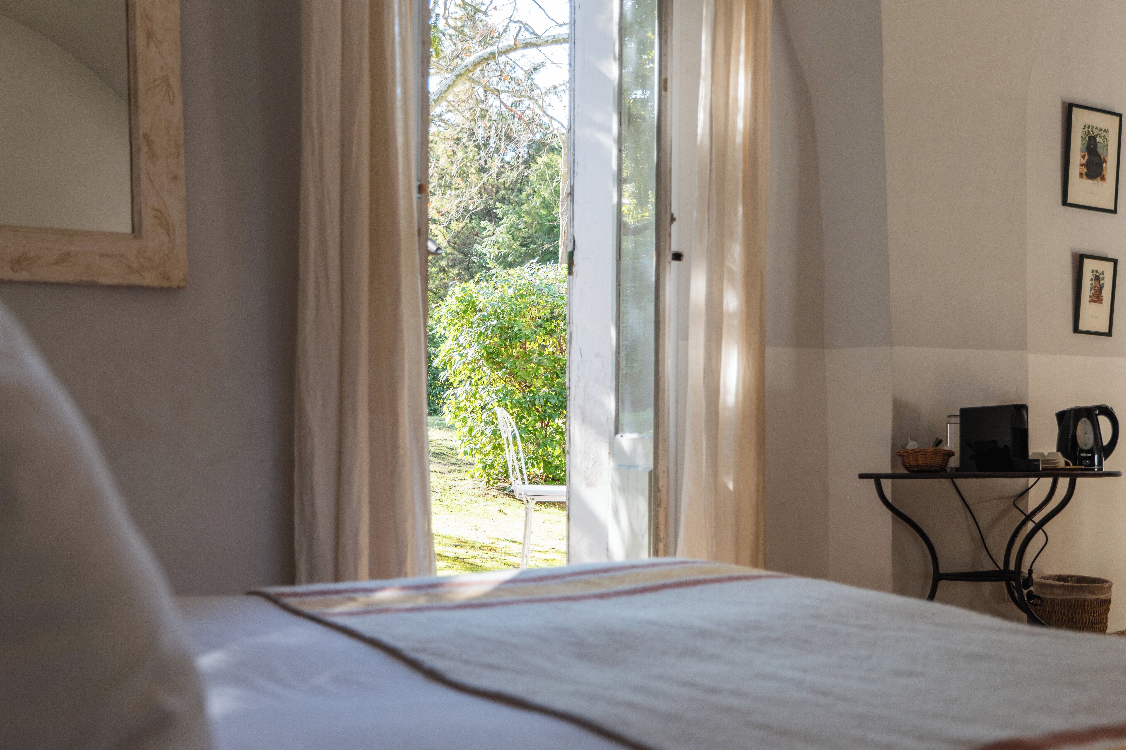 A bedroom with a bed, a window with curtains, a small table with a basket and a kettle, and a framed picture on the wall.
