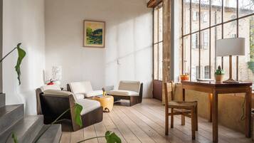 A room with a wooden floor, a sofa, a chair, a table with a lamp, and a window with a view of buildings.