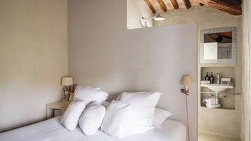 Una camera da letto con un letto, cuscini, un comodino, una lampada, uno specchio e un bagno con un lavandino e un porta asciugamani.