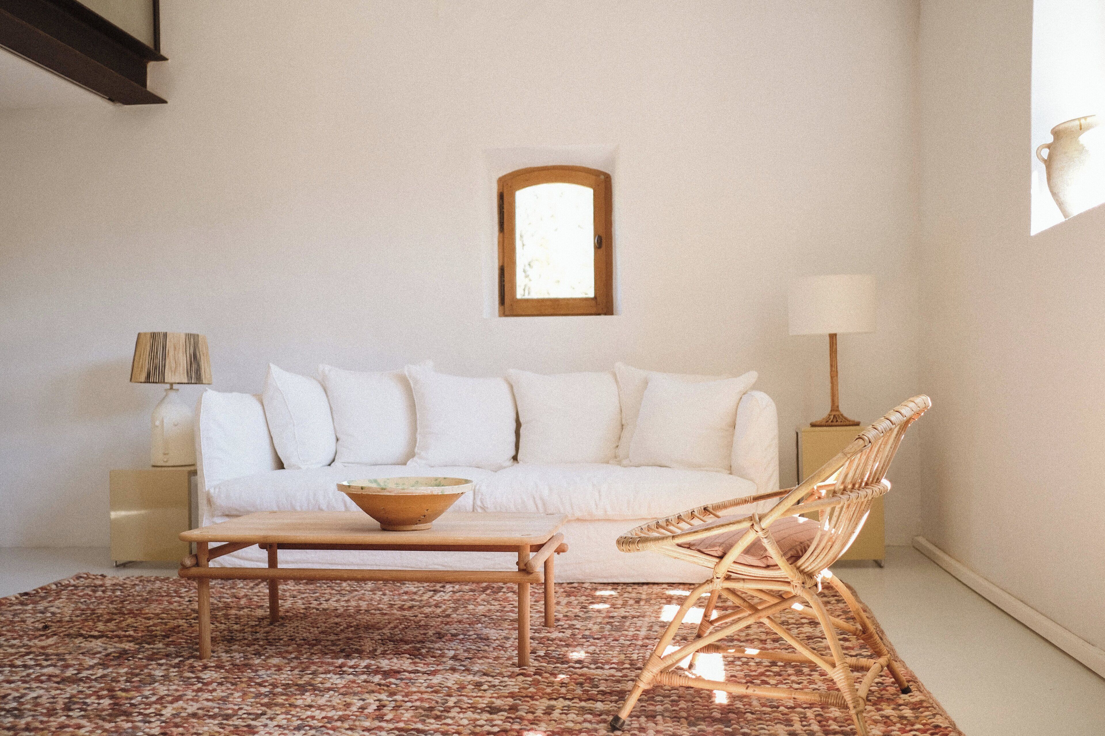 Ett minimalistiskt vardagsrum med en vit soffa, ett träbord, en fåtölj i rotting och en liten bordslampa.