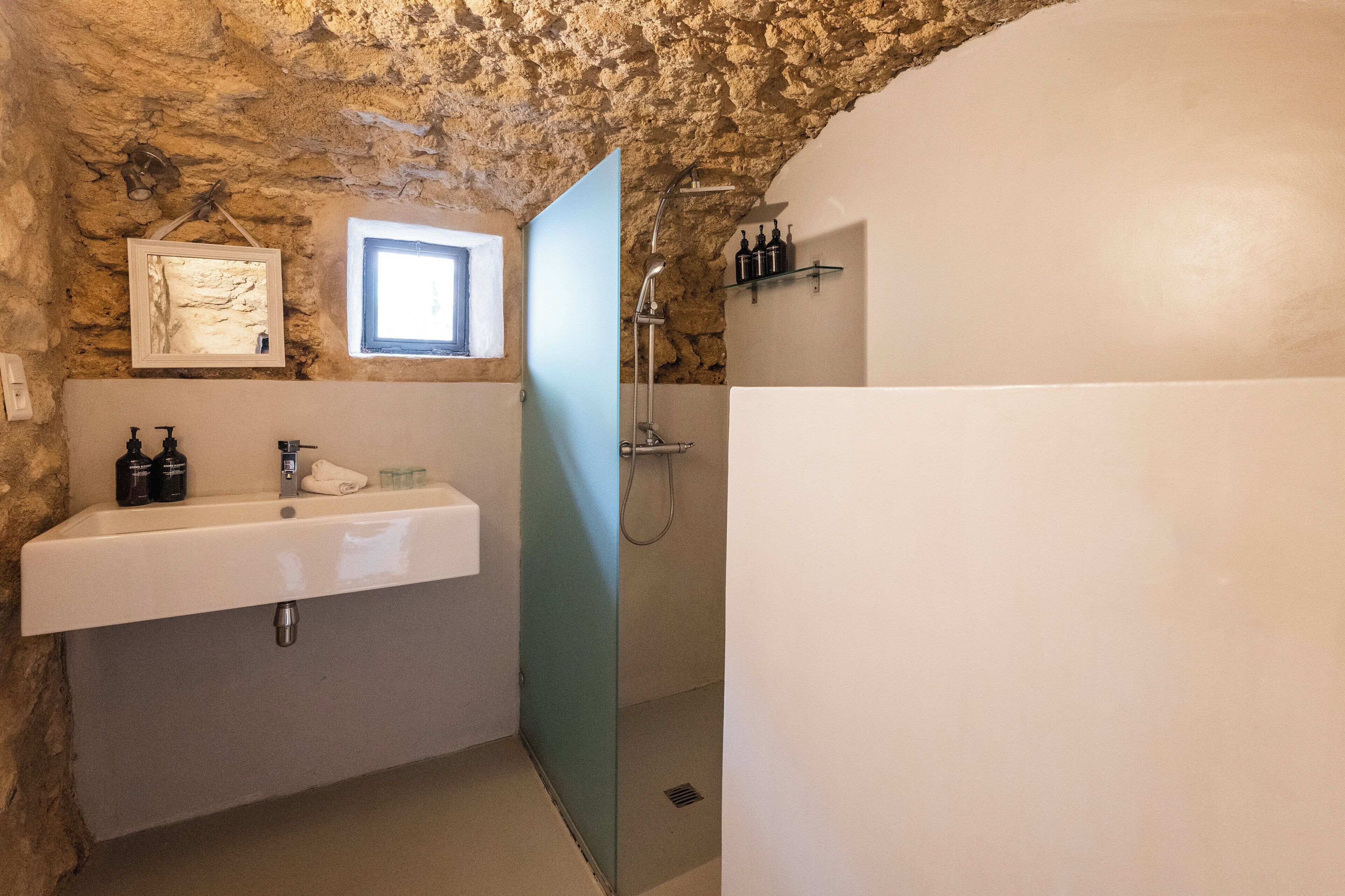 A bathroom with a stone wall, a white sink, a shower, and a small window.