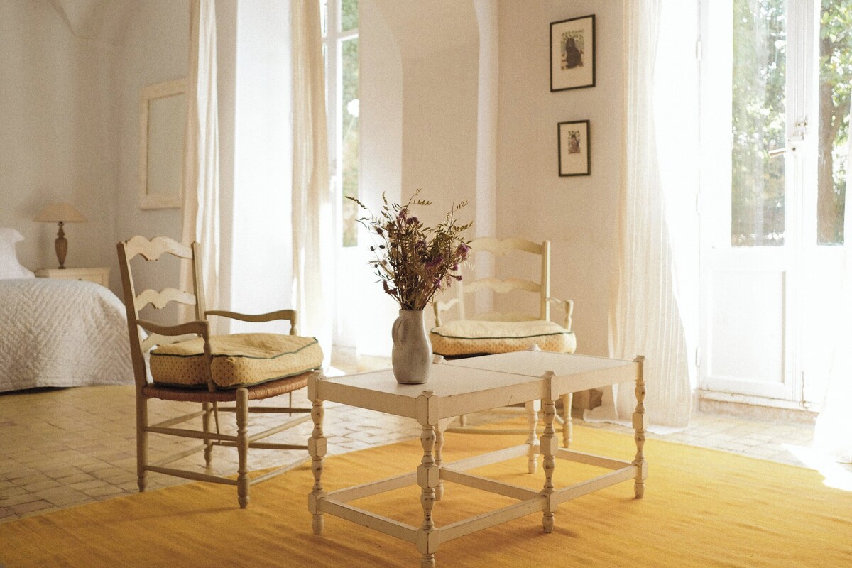 Una habitación con una cama, dos sillas, una mesa y un florero con flores.