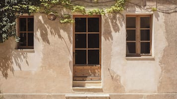 A building with a central door and two windows on either side, a small set of steps leading to the door, and a wall with some greenery growing on it.