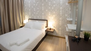 Una cama bien tendida con sábanas blancas, una mesita de noche con lámpara, una planta colgante y un perchero de madera.
