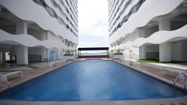 Un área de piscina moderna de hotel, con una piscina central rodeada de balcones blancos y una zona de estar cubierta.