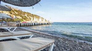 A pebbled beach with lounge chairs and umbrellas, a pier extending into the sea, and buildings on a hillside in the background. - Tasada Otel (Karaburun)