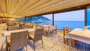 An outdoor dining area with wicker chairs and tables, overlooking a mountainous coastline. - Tasada Otel (Karaburun)