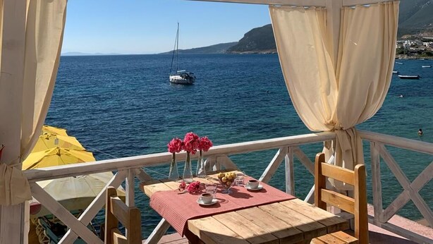 A wooden table with a red tablecloth, a vase with pink flowers, and a bowl of lemons and oranges on a balcony overlooking the sea. - Tasada Otel (Karaburun)