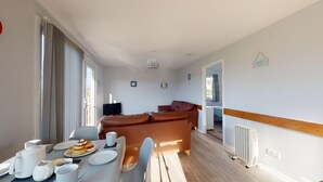 A modern living room with a brown leather sofa, a dining table set with plates and cups, and a television. - A21 Solano, Riviere Towans (Hayle)