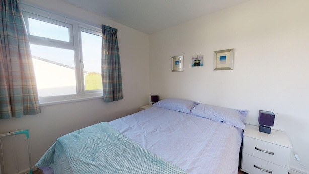 A bedroom with a bed, a bedside table, a lamp, and a window with curtains. - A21 Solano, Riviere Towans (Hayle)