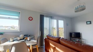 Uma sala de estar com sofá, mesa de jantar com cadeiras, televisão e janela com cortinas.