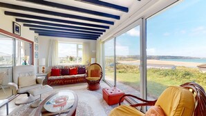 Ein geräumiges Wohnzimmer mit einer Couch, Sesseln und einem Couchtisch, große Fenster mit Blick auf Strand und Hügel.