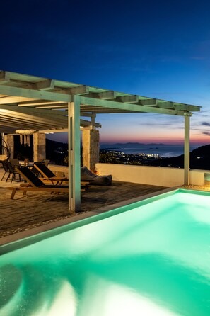 An outdoor pool area with lounge chairs and a pergola, overlooking a cityscape at dusk. - Paros Nereids Villas (Paros)