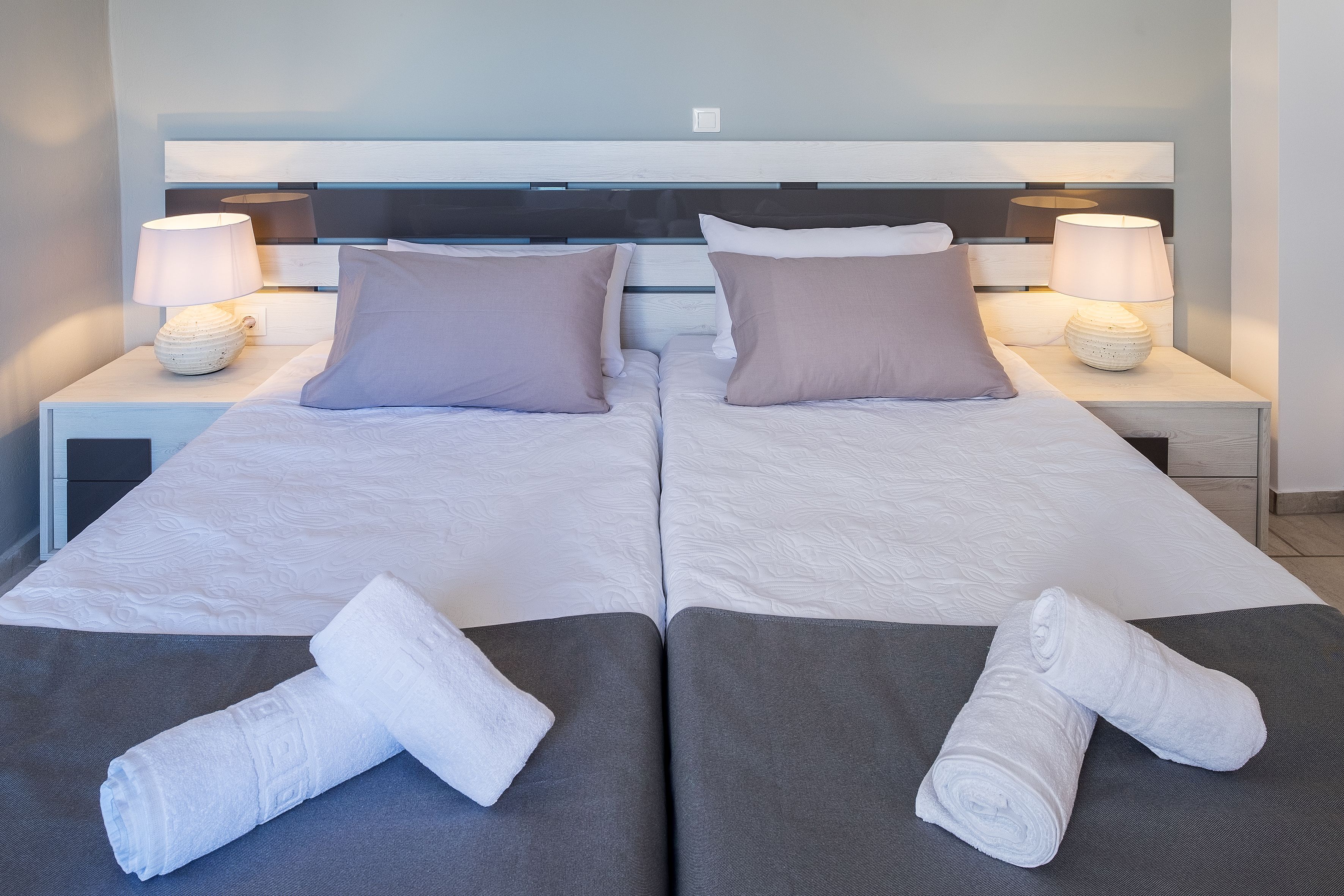 A double bed with two pillows, a nightstand with a lamp, and rolled towels on the bed.