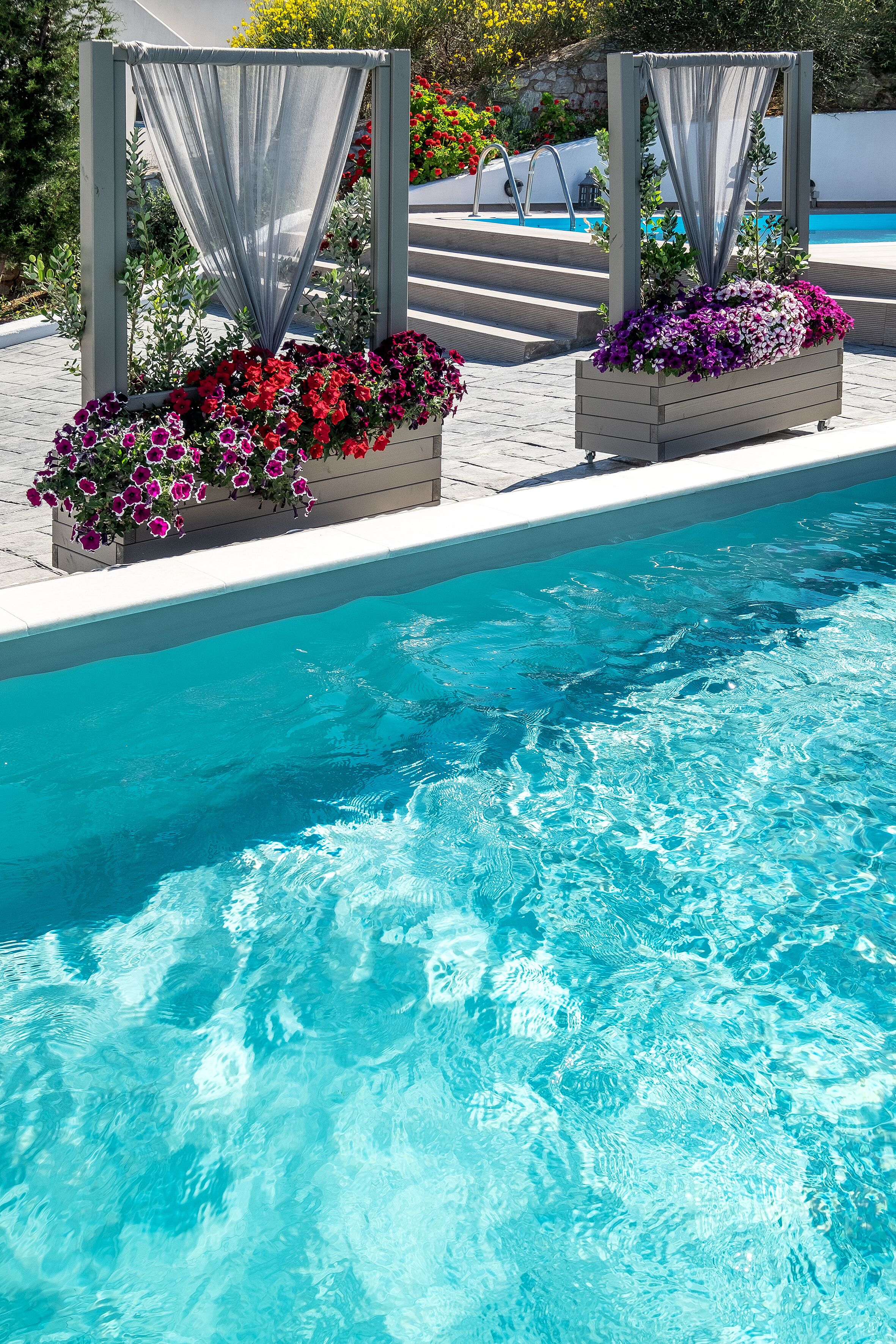 A swimming pool with clear blue water, surrounded by flower planters with red and purple flowers, and a covered seating area with a poolside view.