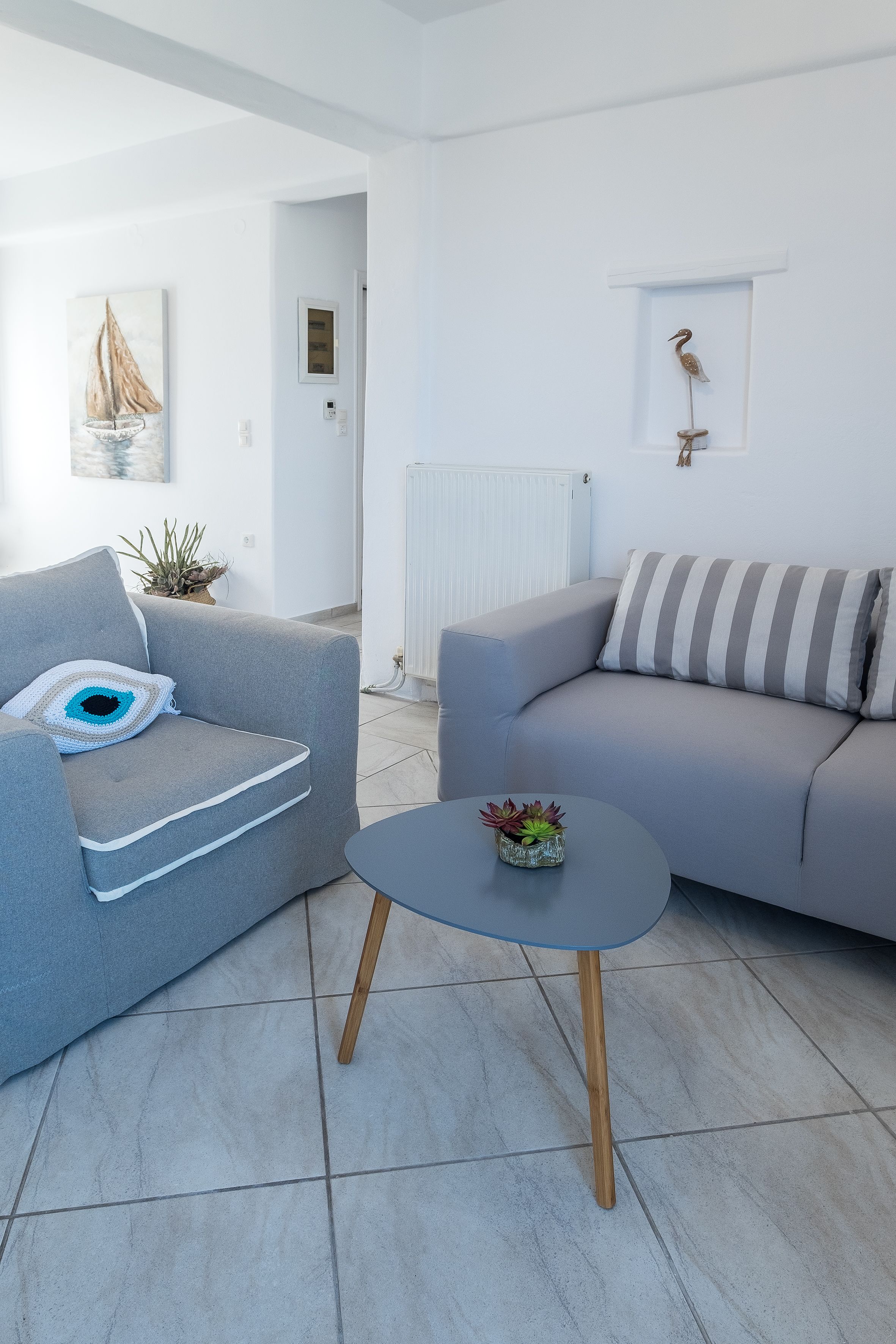Een moderne woonkamer met een grijze bank, een klein rond tafeltje met een plant, en een aan de muur bevestigde vogelsculptuur.
