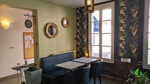 Een restaurantinterieur met een groene wand, twee klokken, een prikbord, een zithoek met een bank en tafels, en een raam met uitzicht op de straat.