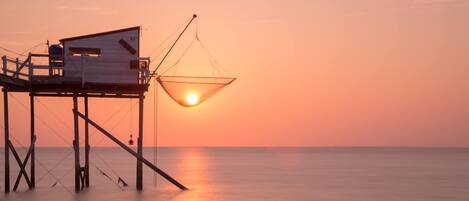 A wooden pier with a fishing hut and a hanging net, overlooking a calm sea at sunset.