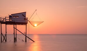 Une jetée en bois, avec une cabane de pêche et un filet suspendu, surplombant une mer calme au coucher du soleil.