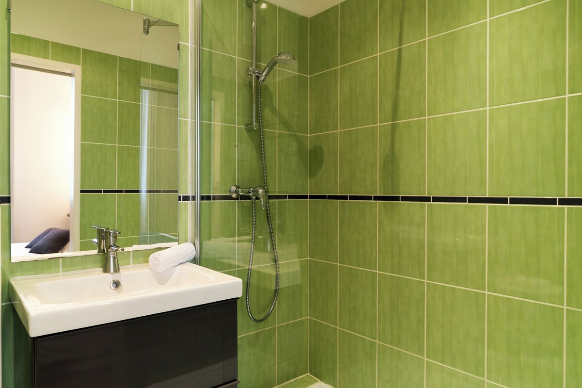 Un baño con azulejos verdes, un lavamanos blanco y una zona de ducha con mampara de vidrio.