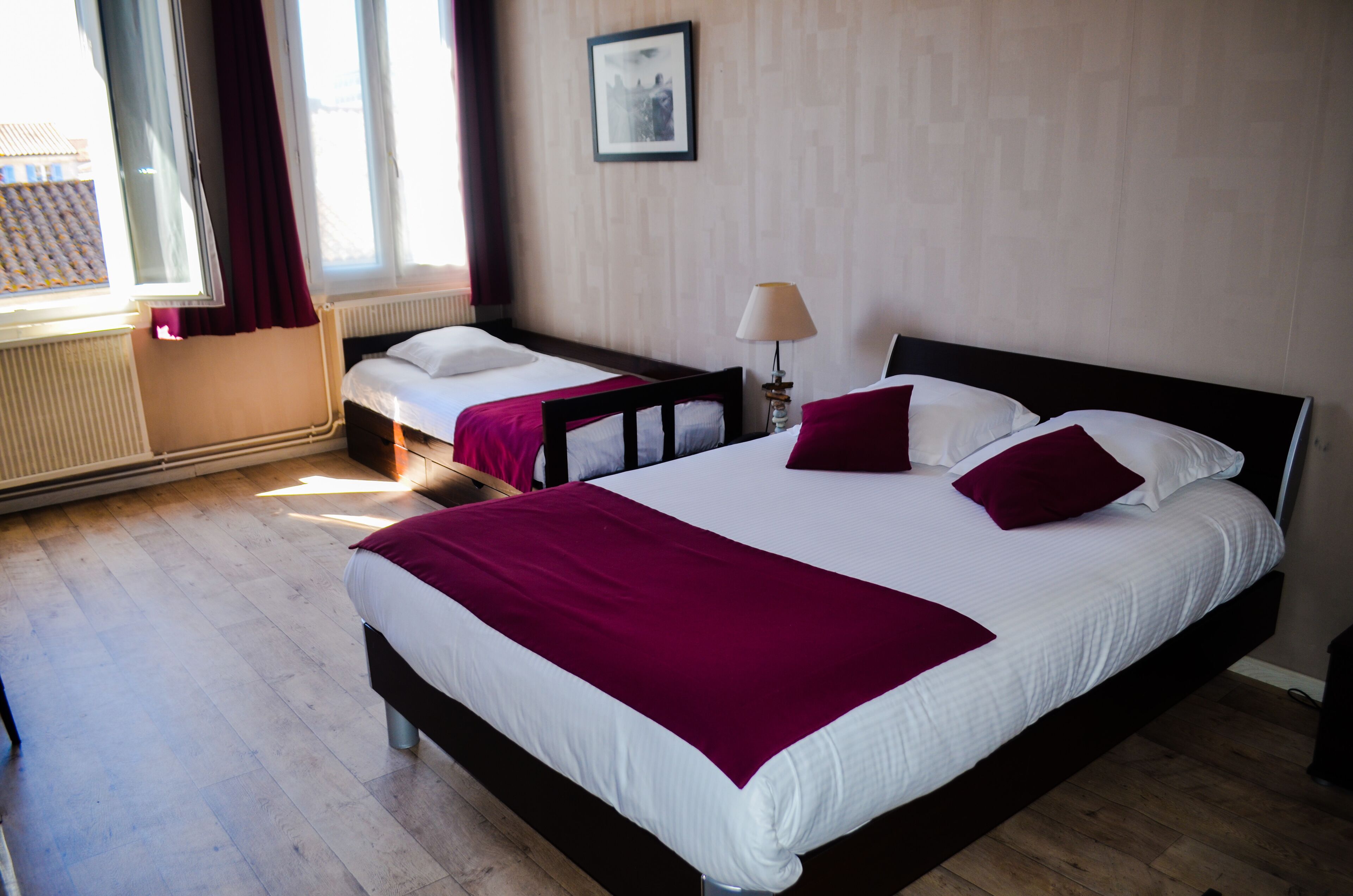 Ett hotellrum med två sängar, ett stort fönster med gardiner, en inramad tavla på väggen och en sänglampa.