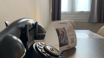 Un vieux téléphone noir posé sur une table en bois, avec une lampe et une brochure en arrière-plan.