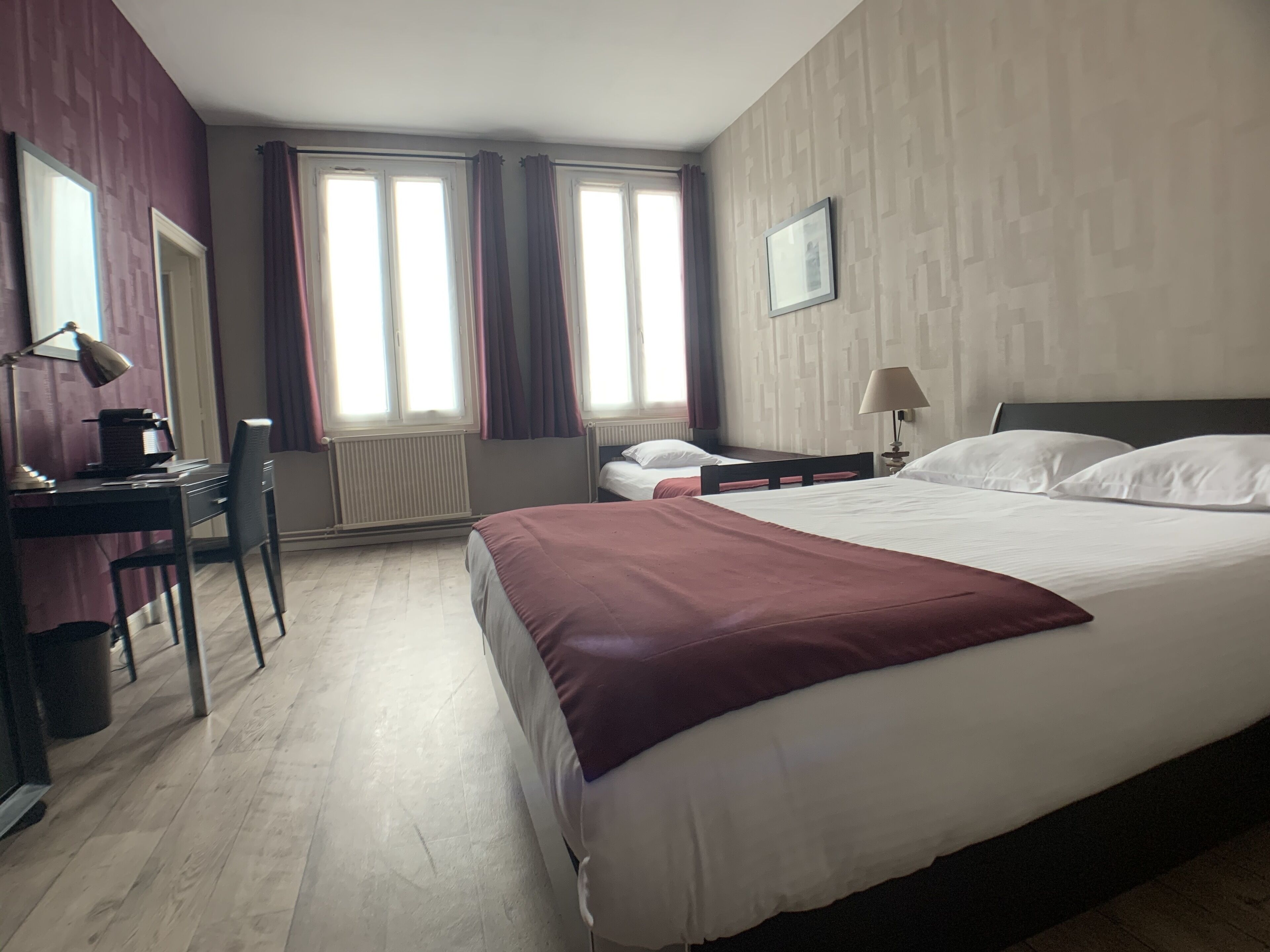 Habitación de hotel con una cama grande, un escritorio con silla, un radiador y dos ventanas con cortinas.