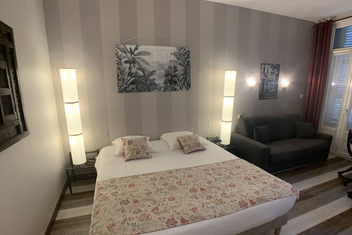 Habitación de hotel con una cama grande, dos lámparas de noche, un sofá, un cuadro enmarcado en la pared y una ventana con cortinas.