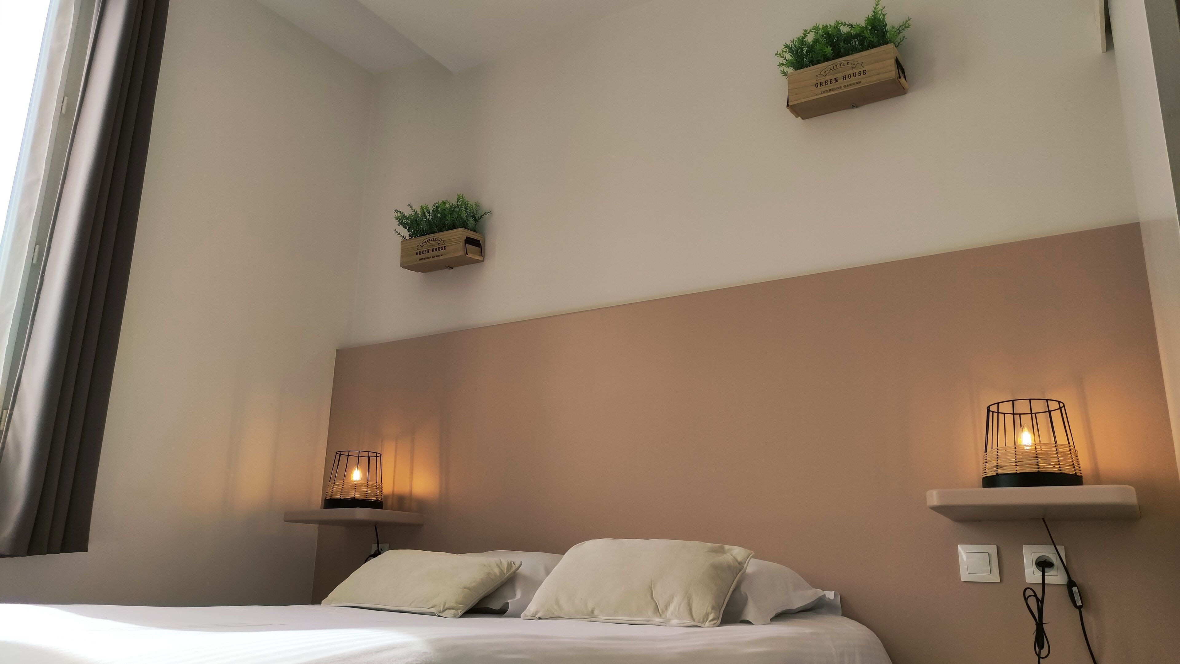Un dormitorio con una cama, dos mesitas de noche con lámparas, un estante en la pared con plantas, y un enchufe en la pared.