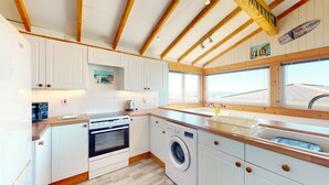 A kitchen with white cabinets, wooden beams, a stove, a washing machine, and a window with a view. - F46 Holiday Haunt, Riviere Towans (HAYLE)