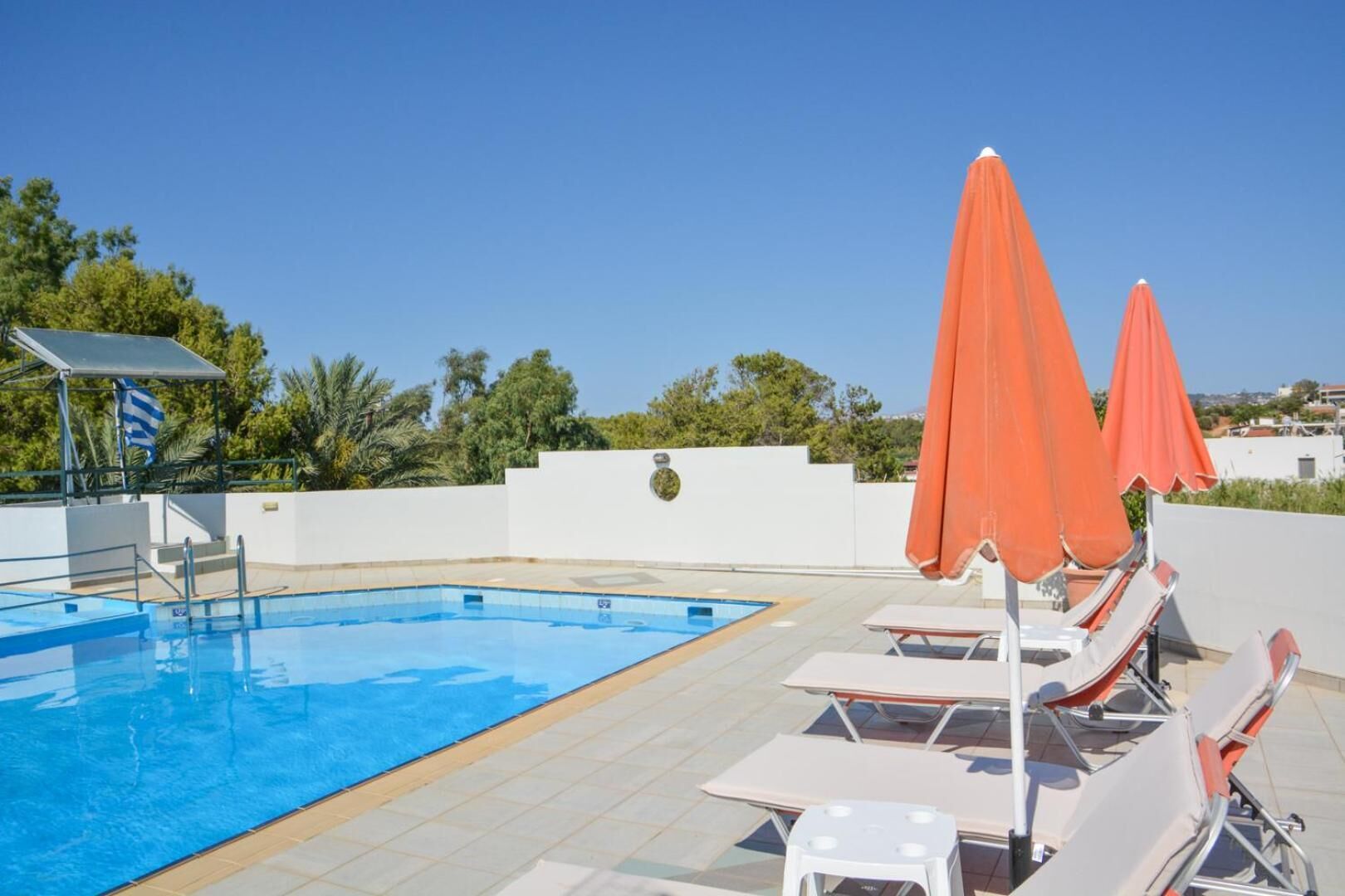 En swimmingpool med liggestole og orange parasoller, omgivet af en hvid mur og grønt.