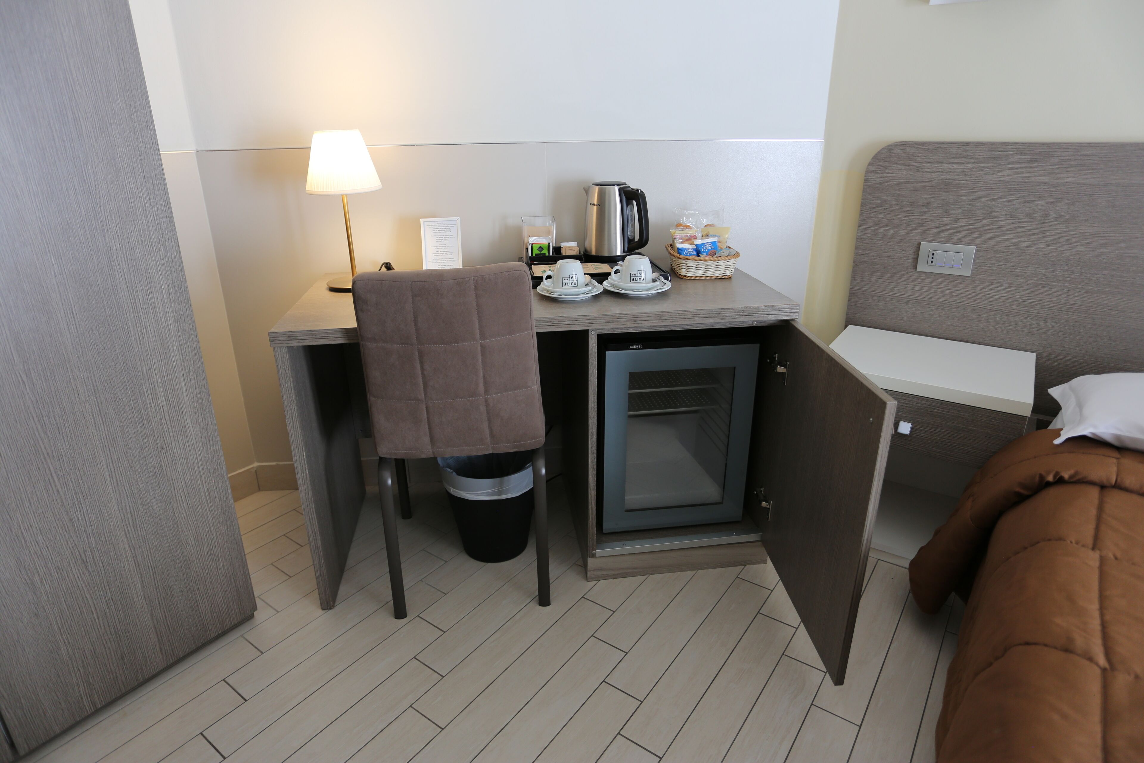 Et lille hotelværelse med tekøkken, et lille køleskab, et skrivebord med lampe samt en seng med brunt lagen.
