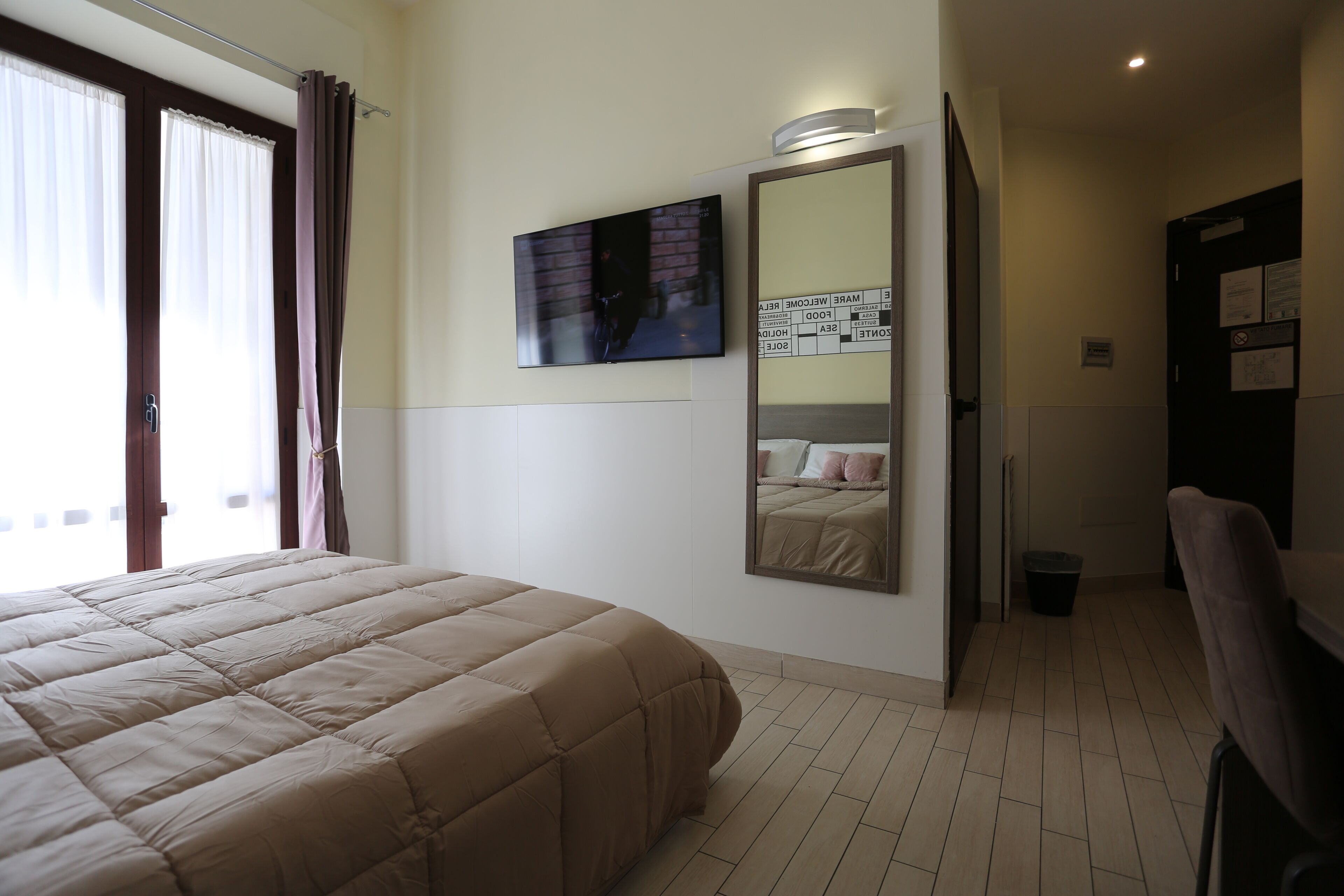 A hotel room with a large bed, a flat-screen TV mounted on the wall, a mirror, and a door with a sign.