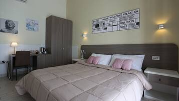 A hotel room with a bed, a desk, a chair, a wardrobe, and a wall sign listing amenities.