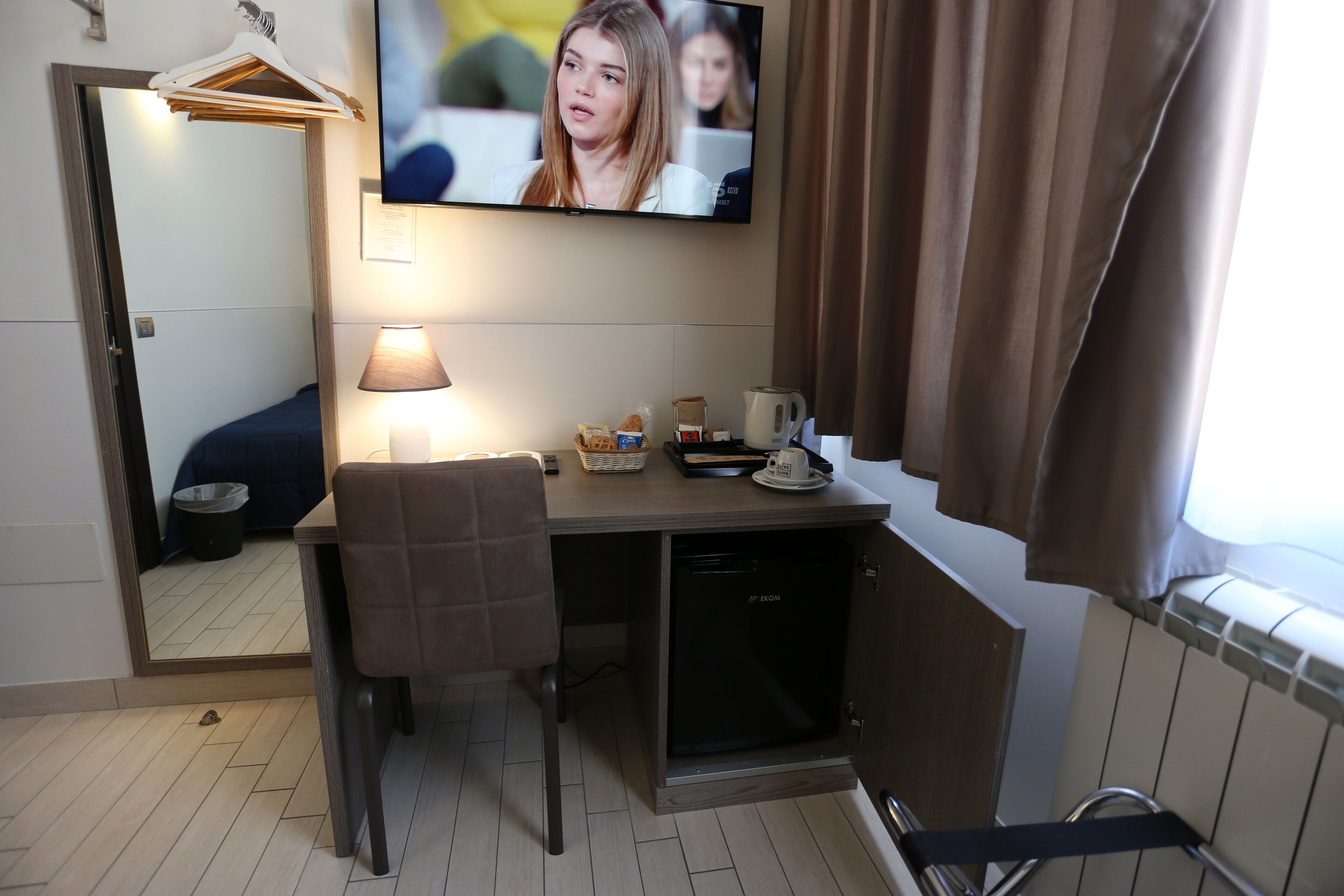 Habitación de hotel con escritorio, silla, televisor, lámpara y una pequeña zona de kitchenette.