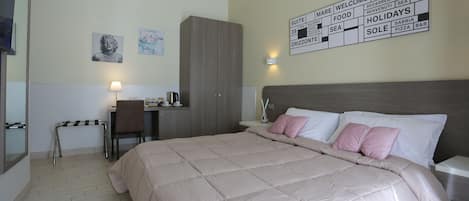 A hotel room with a bed, a desk, a chair, and a wardrobe. There is a wall sign with various amenities listed.