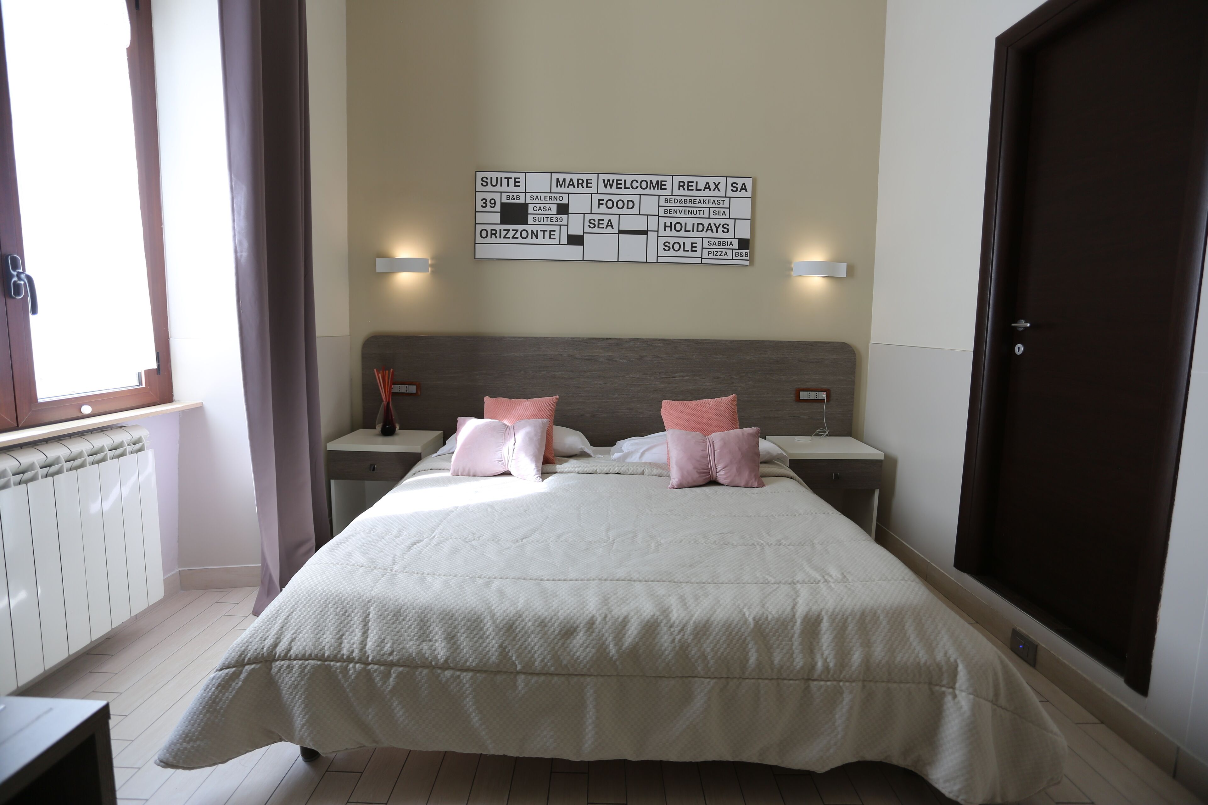 Ein Hotelzimmer mit Bett, zwei Nachttischen, einem an der Wand befestigten Schild mit Informationen zu den Zimmerausstattungen, einem Fenster mit Vorhängen und einer Tür.