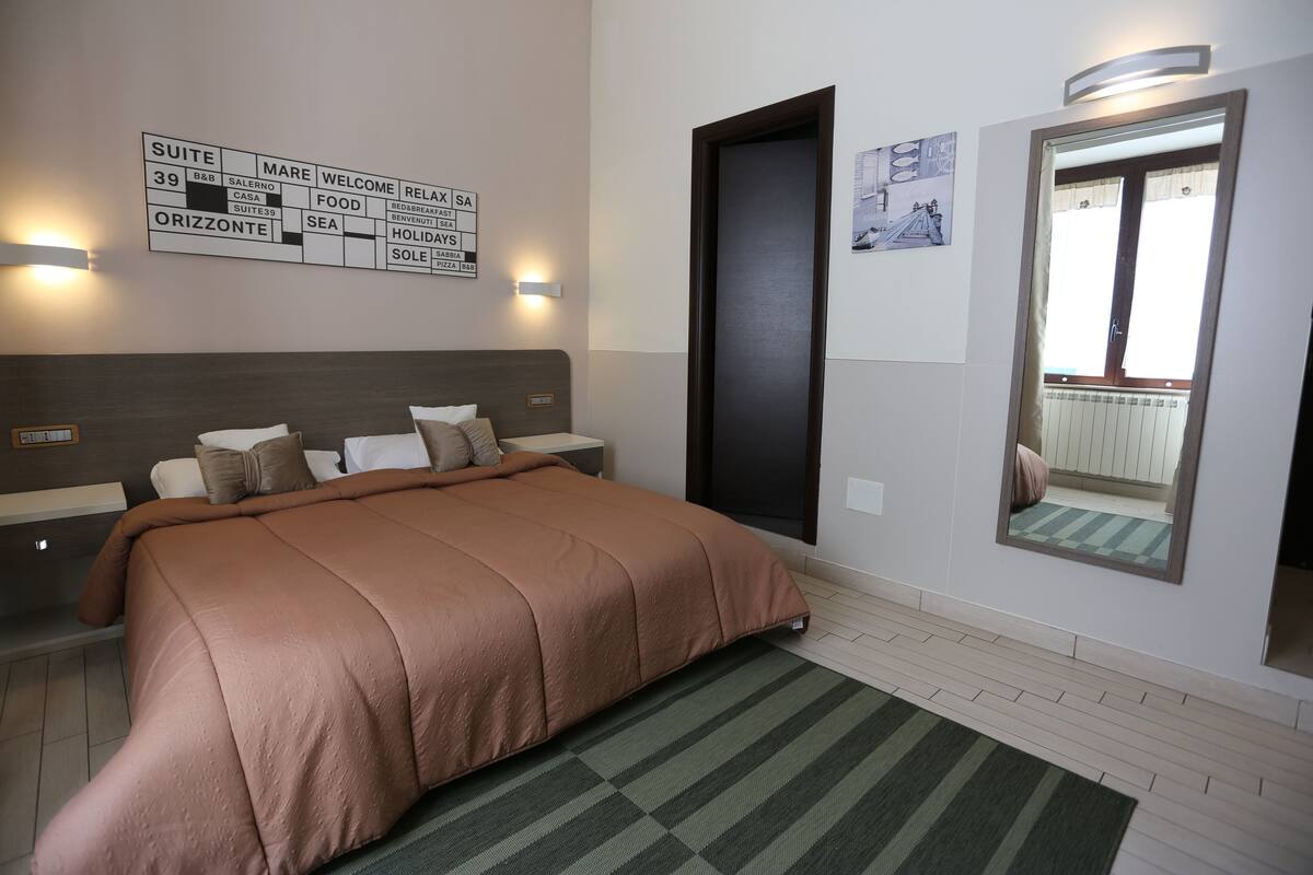 A hotel room with a large bed, a nightstand, a wall-mounted sign with room information, a mirror, and a view of the outside balcony.